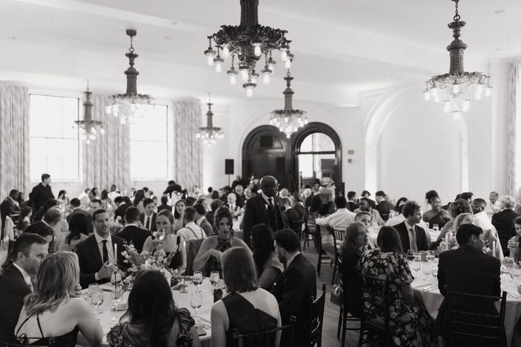 Black-and-white view of wedding reception hall filled with guests, chandeliers overhead and tables styled with cohesive wedding flowers