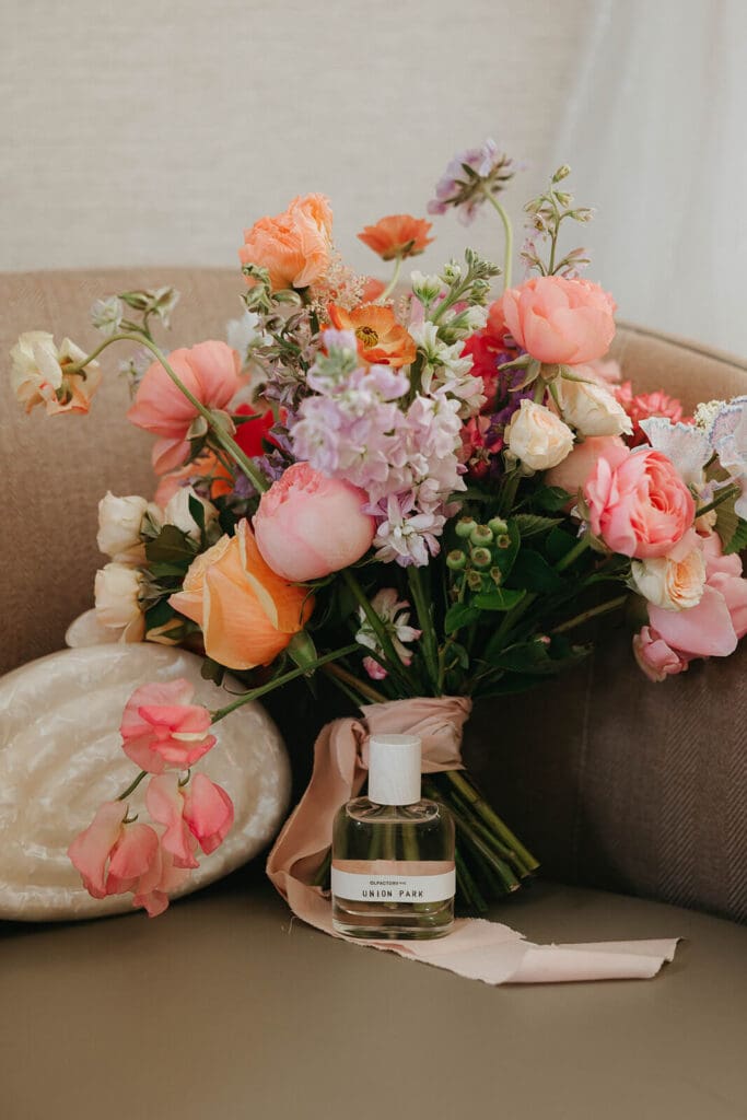 Wedding bouquet in blush, coral, and lilac tones placed beside perfume and ribbon on a velvet chair