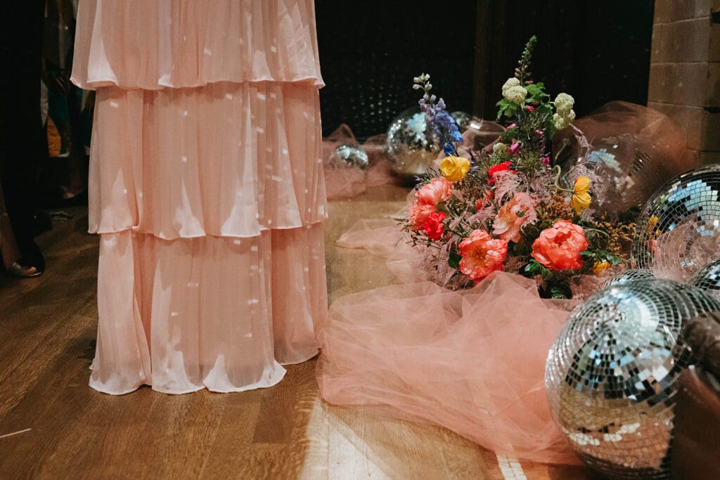 Guest in a pink tiered dress standing beside bold cohesive wedding flowers and reflective disco balls at a reception entrance