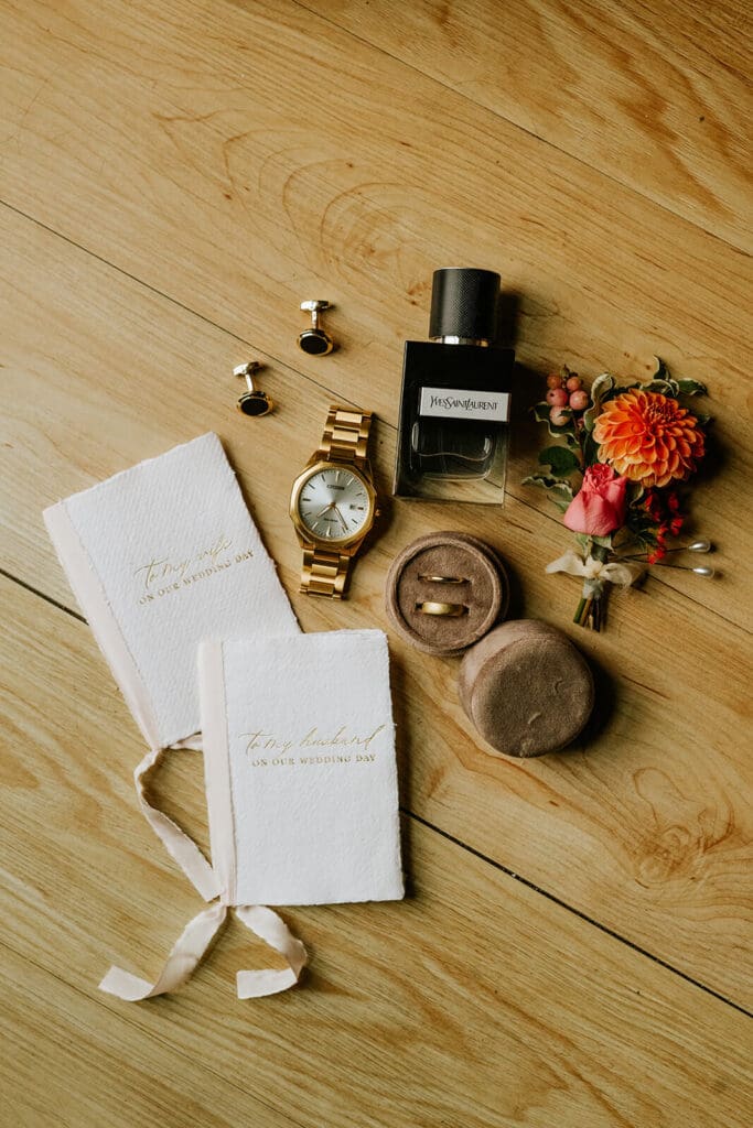 Groom’s flatlay with vow books, watch, cologne, cufflinks, and boutonnière styled on a wood floor.