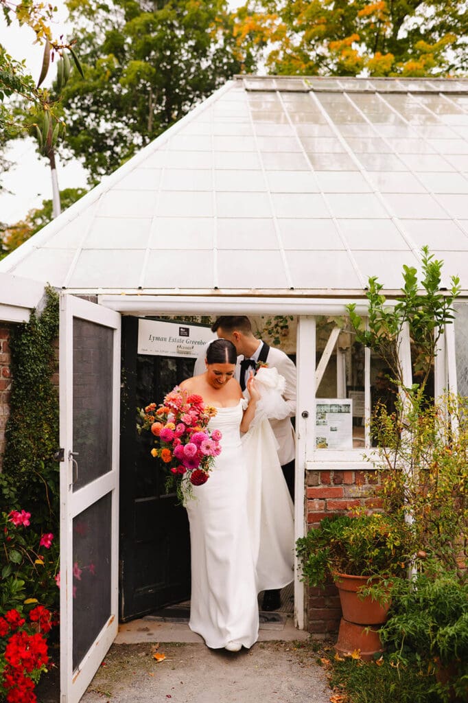 Bride and groom exiting the Lyman Estate greenhouse, framed by lush plants and carrying a bright seasonal bouquet of summer wedding flowers