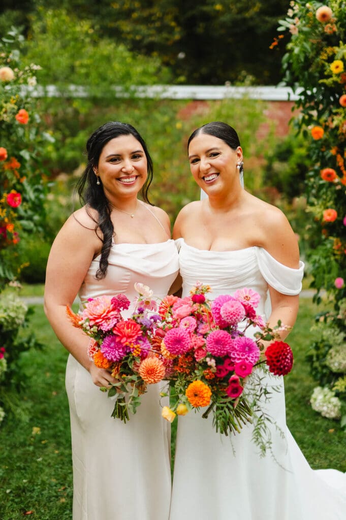 Bride and bridesmaid smiling side by side, holding matching bouquets full of zinnias and dahlias in saturated summer hues.