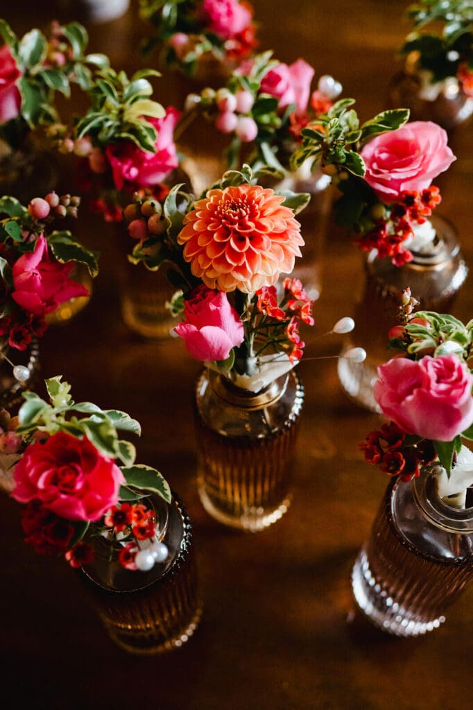 Collection of bud vases with coral dahlias, pink spray roses, and summer greenery arranged as table decor.