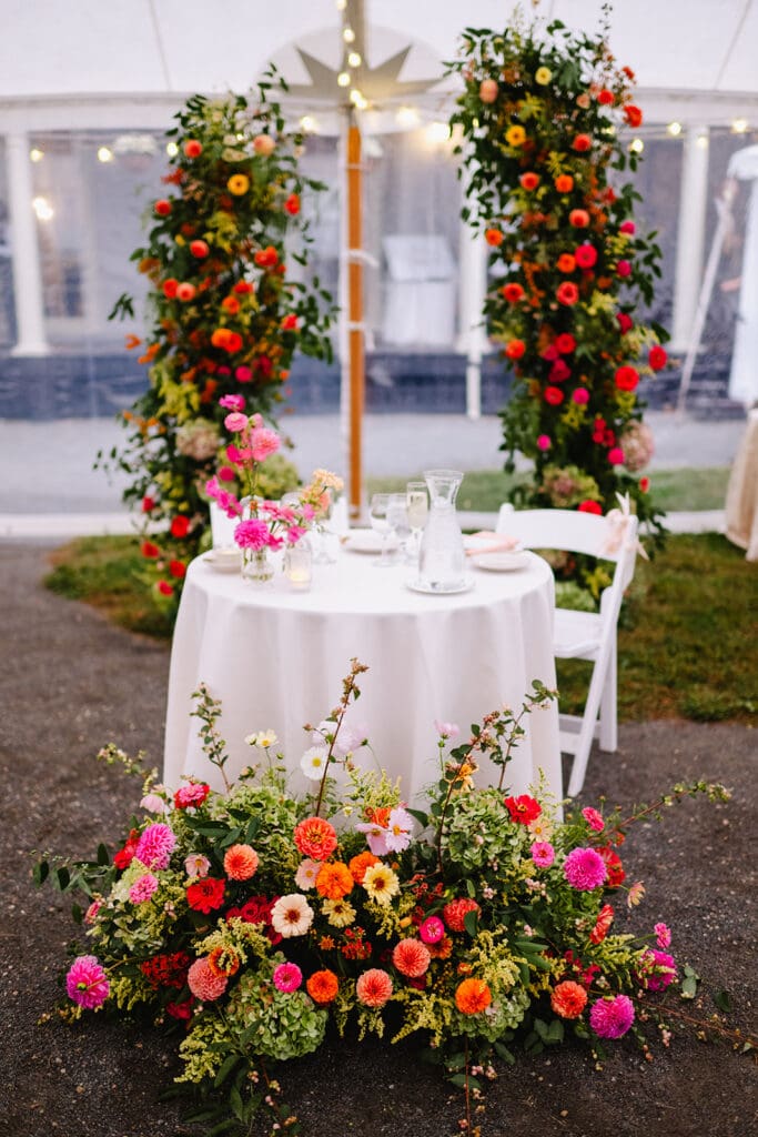 Sweetheart table framed by a garden-style floor arrangement and floral pillars under a sailcloth tent.