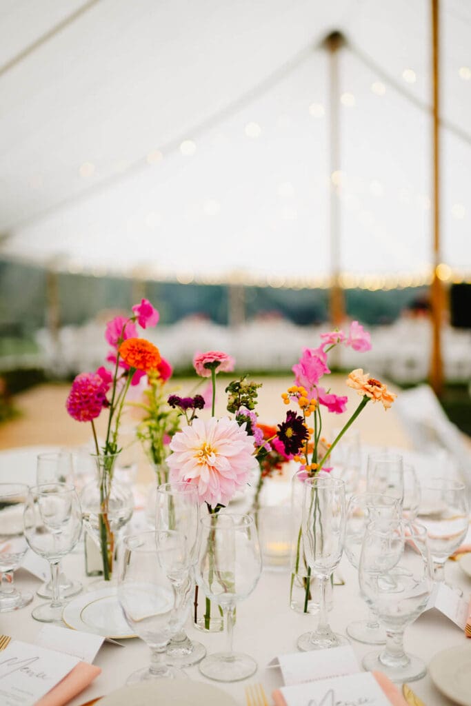 Tent reception table with a loose arrangement of summer wedding flowers in glass bud vases and soft candlelight.