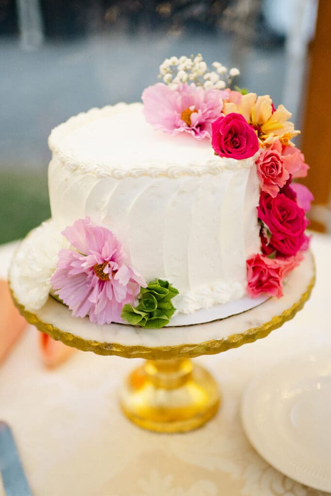 White buttercream wedding cake topped with fresh blooms in pink and blush tones, displayed on a gold stand.