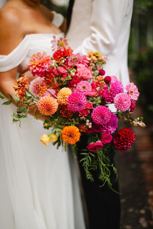 Close-up of bride holding a bold bouquet of magenta, coral, orange, and pink summer wedding flowers against her off-the-shoulder gown.