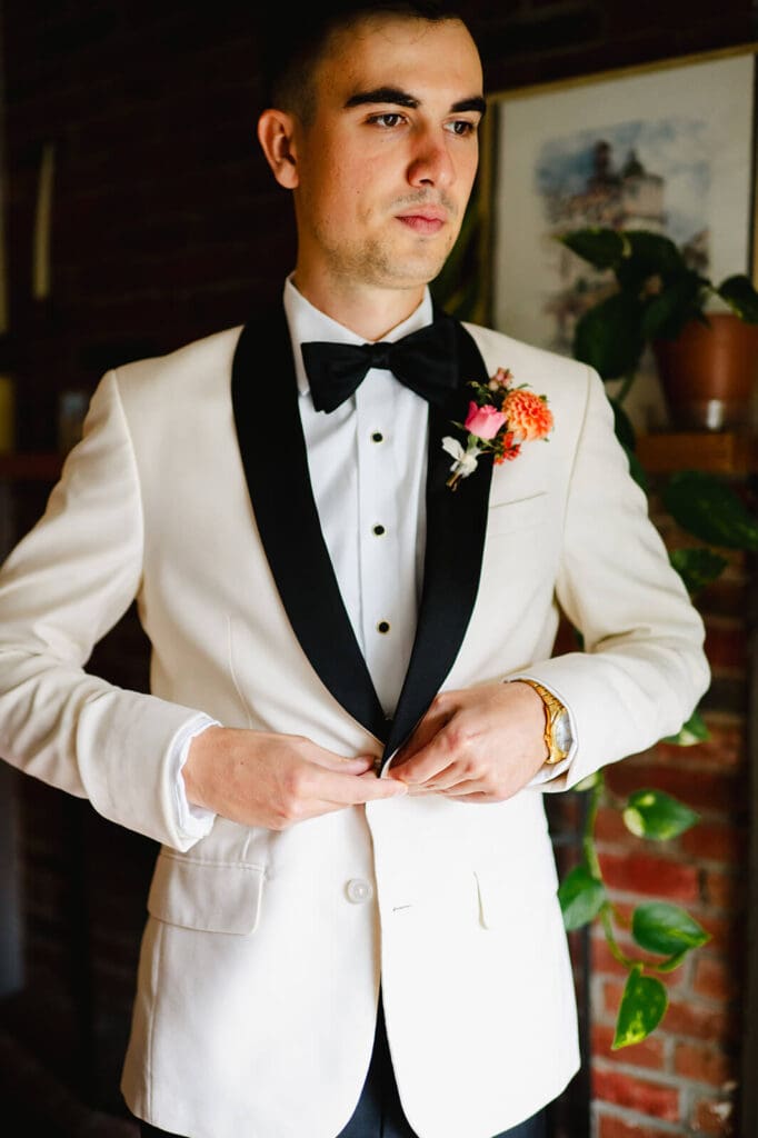Groom fastening his white tuxedo jacket with a peach boutonnière, standing in front of a brick wall and houseplants.