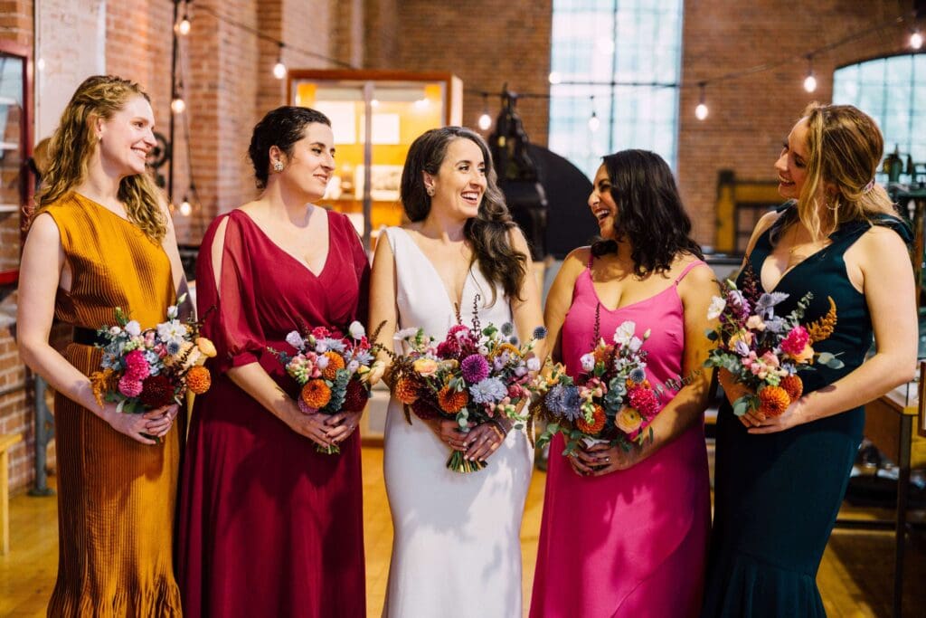 Bridesmaids in mustard, crimson, and magenta dresses holding lush fall floral bouquets