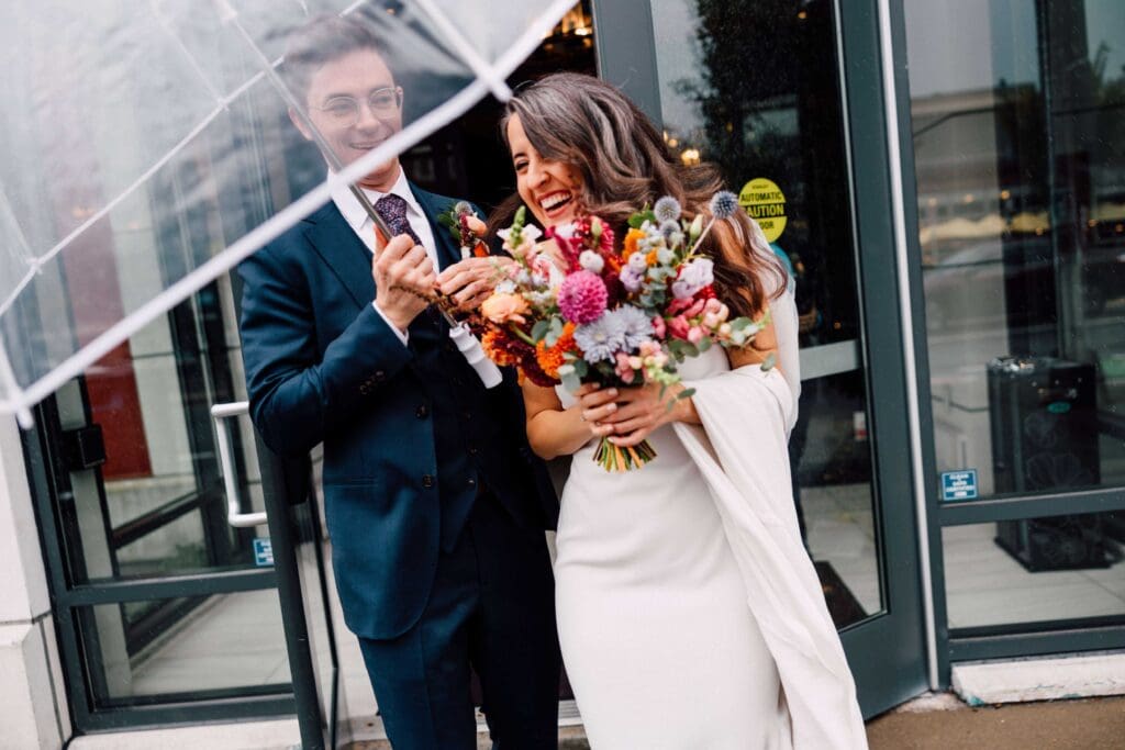 Bride and groom smiling with a vibrant fall bouquet outside a modern Boston venue

