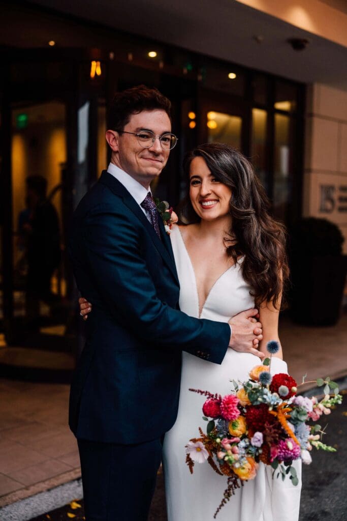 Newlyweds smiling in front of hotel entryway with a vibrant fall bouquet