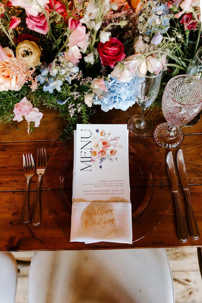 Barn wedding reception table with colorful seasonal flowers and printed menu card styled with glassware.
