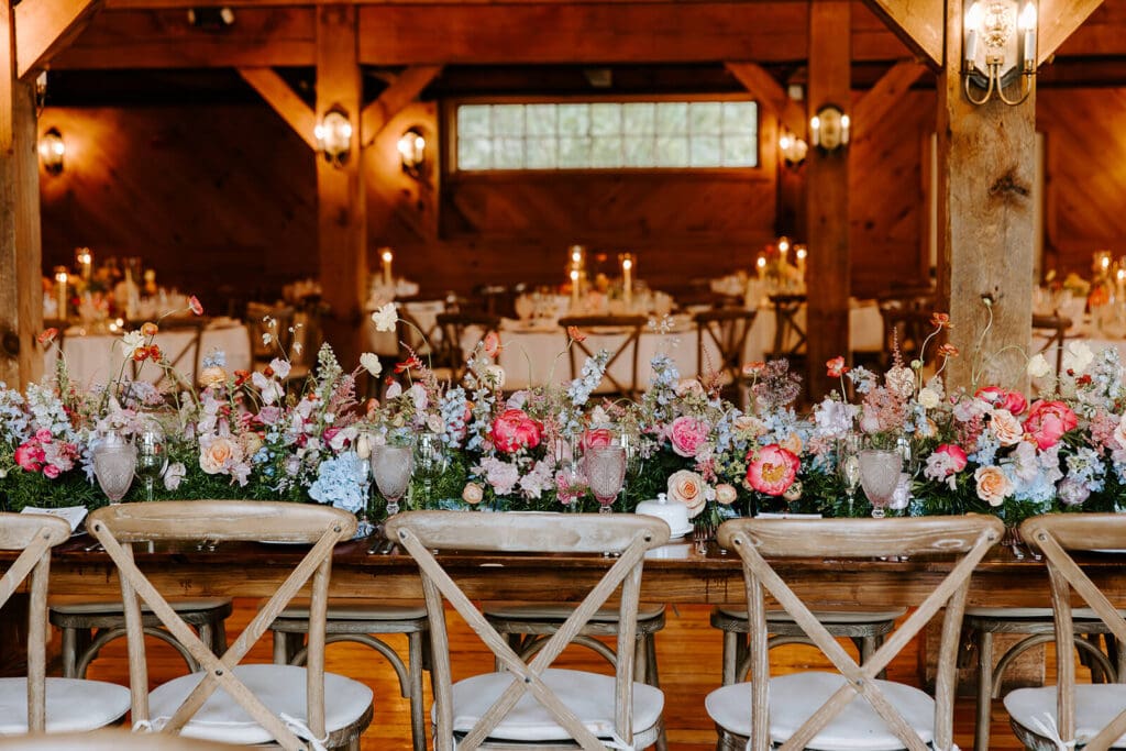 Long head table at a barn wedding venue covered in rustic wedding flowers in pink, peach, and blue tones.