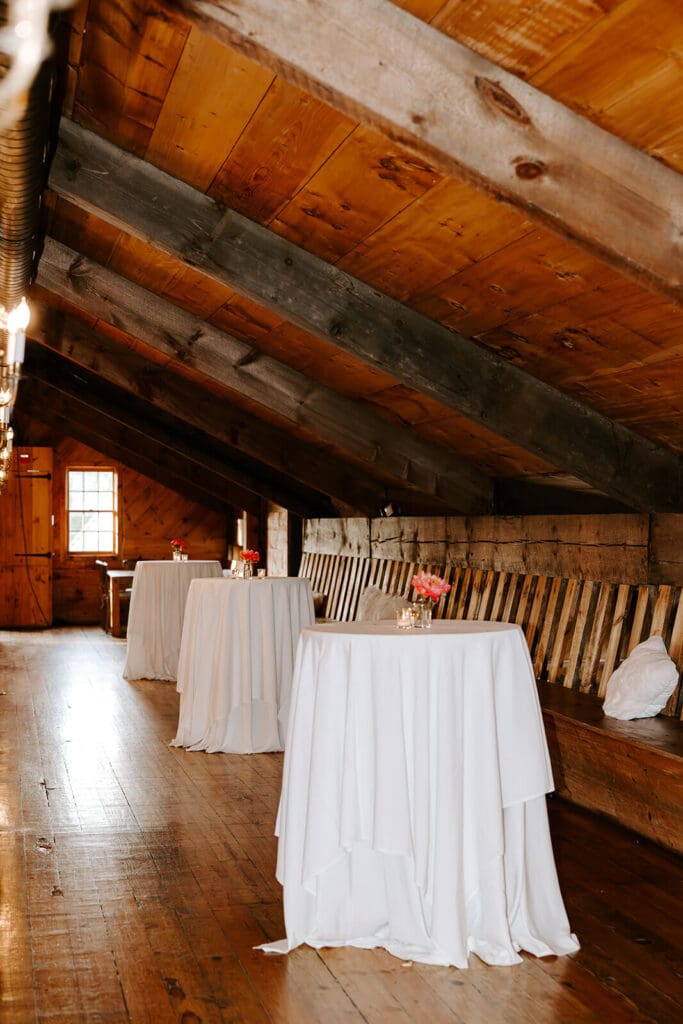Cocktail hour setup at barn wedding venue with high-top tables and rustic wooden beams.