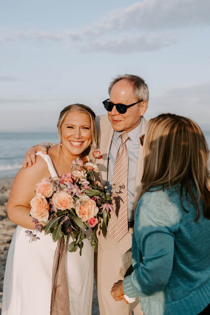 Bride with seasonal bouquet smiling with her father at seaside ceremony before barn wedding reception.