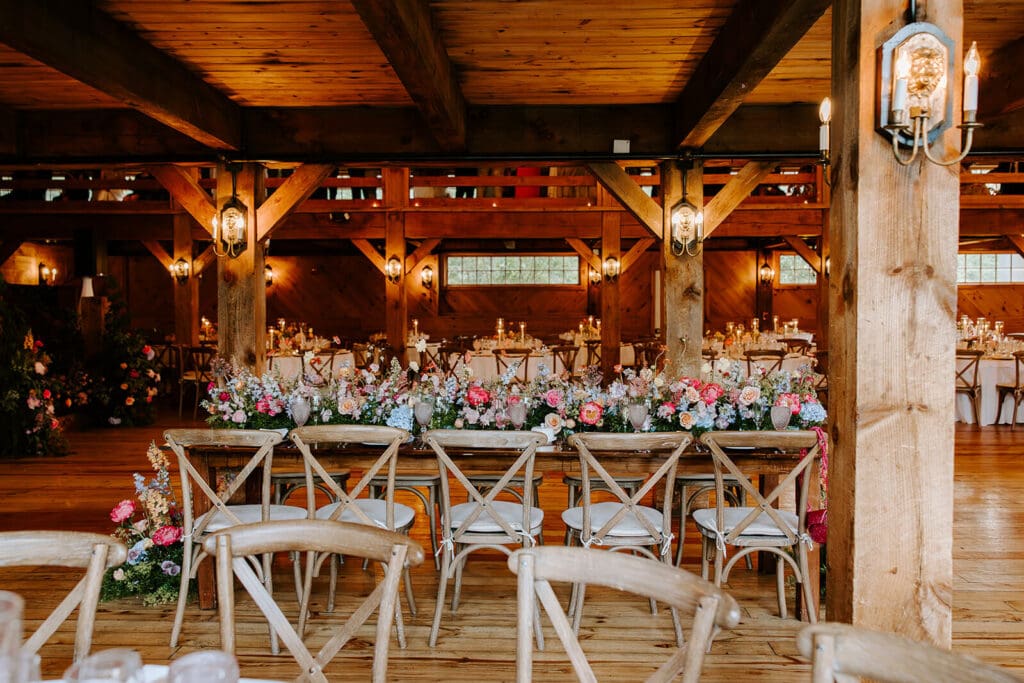 Long rustic head table styled with seasonal flowers in pink, peach, and blue at barn wedding reception.