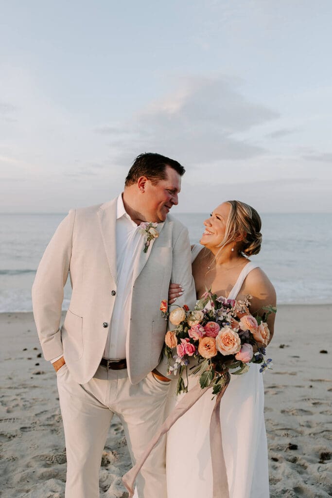 Groom in light suit and bride holding seasonal bouquet of rustic wedding flowers walking along Rexhame Beach.
