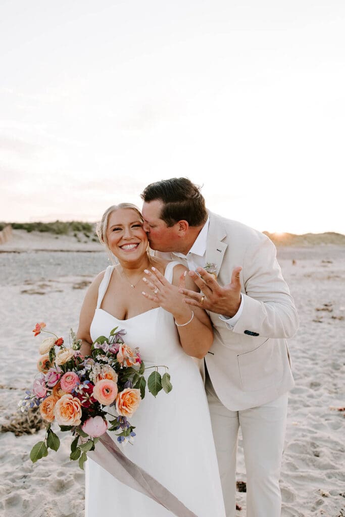 Bride and groom laughing on Rexhame Beach at sunset, holding a bouquet of rustic wedding flowers with seasonal peonies, ranunculus, and garden roses.