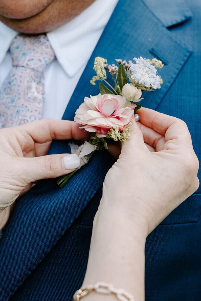 Groom’s boutonnière of seasonal flowers being pinned on blue suit jacket before barn wedding ceremony.
