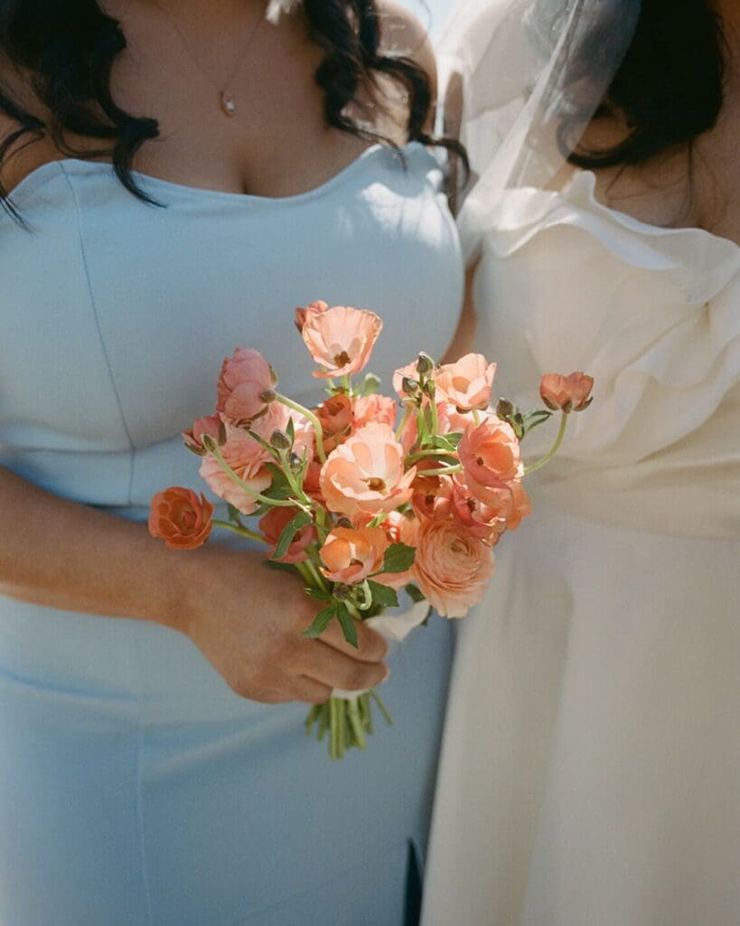 Bridesmaid in light blue dress holding a simple bouquet of coral ranunculus, vibrant seasonal flowers chosen for a May Beauport Hotel Gloucester wedding.