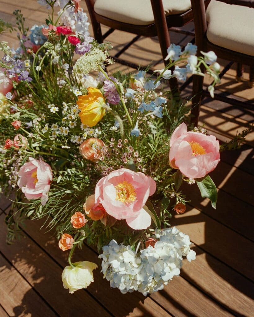Lush garden-style arrangement of colorful wedding flowers with pink peonies, yellow poppies, and blue hydrangea lining the ceremony aisle at an oceanfront Gloucester wedding.