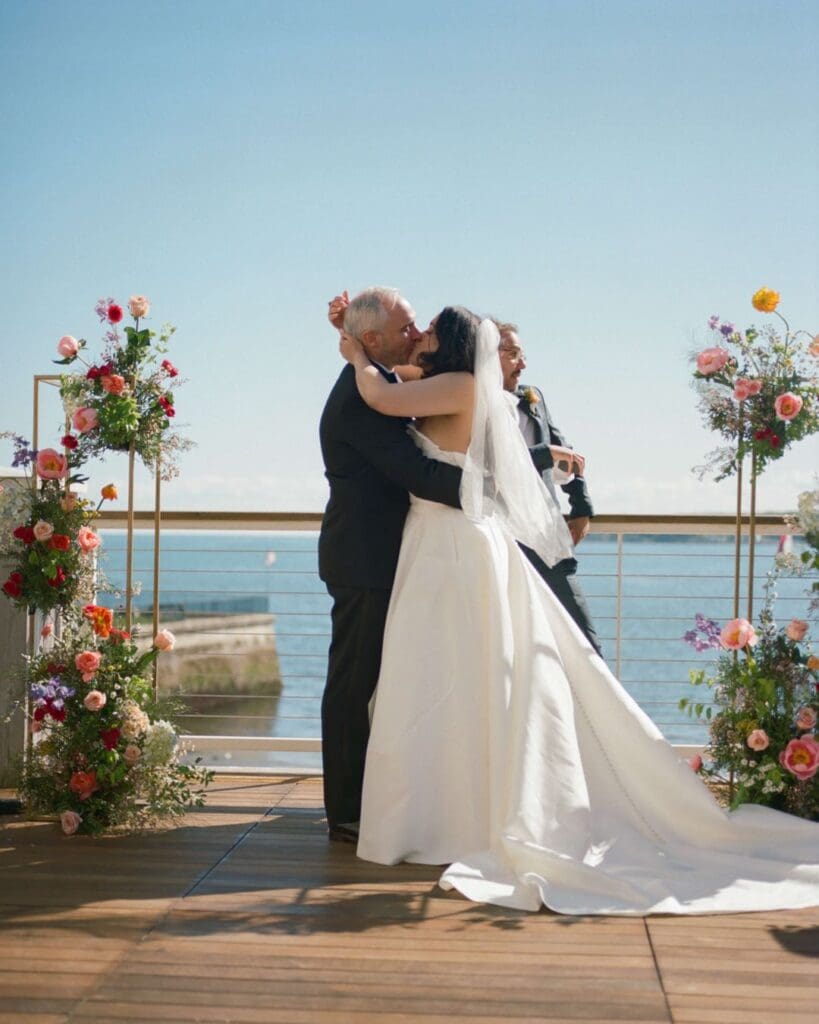 Bride and groom’s first kiss framed by colorful beach wedding flowers, with the Gloucester coastline as a backdrop at the Beauport Hotel.