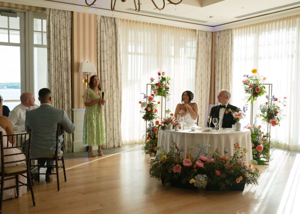 Bride and groom at their sweetheart table during a Beauport Hotel Gloucester wedding, surrounded by colorful May wedding flowers while a guest gives a toast.