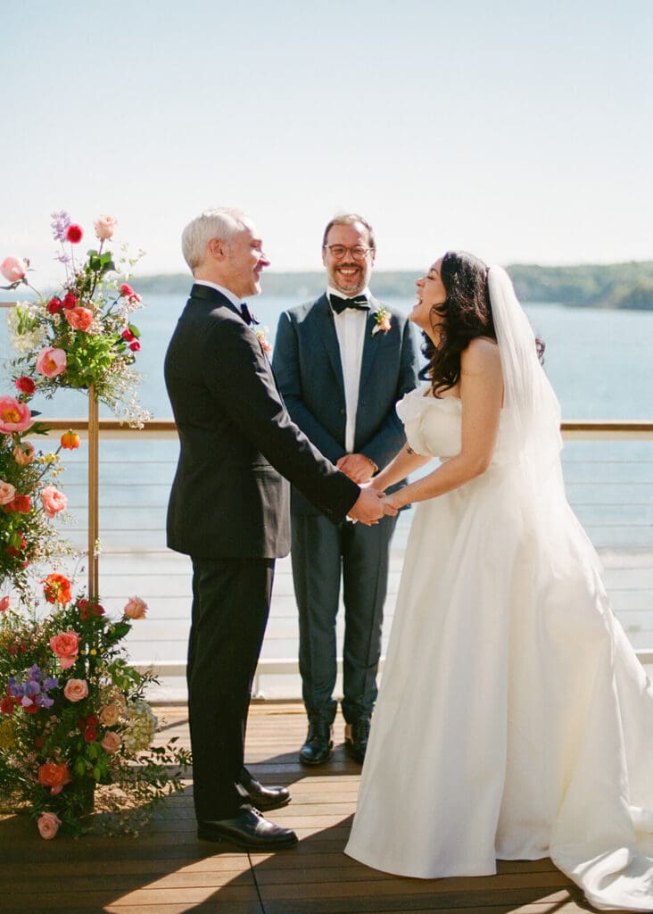Bride and groom exchange vows on the Beauport Hotel deck in Gloucester, surrounded by colorful seasonal flowers for a May ocean wedding ceremony.