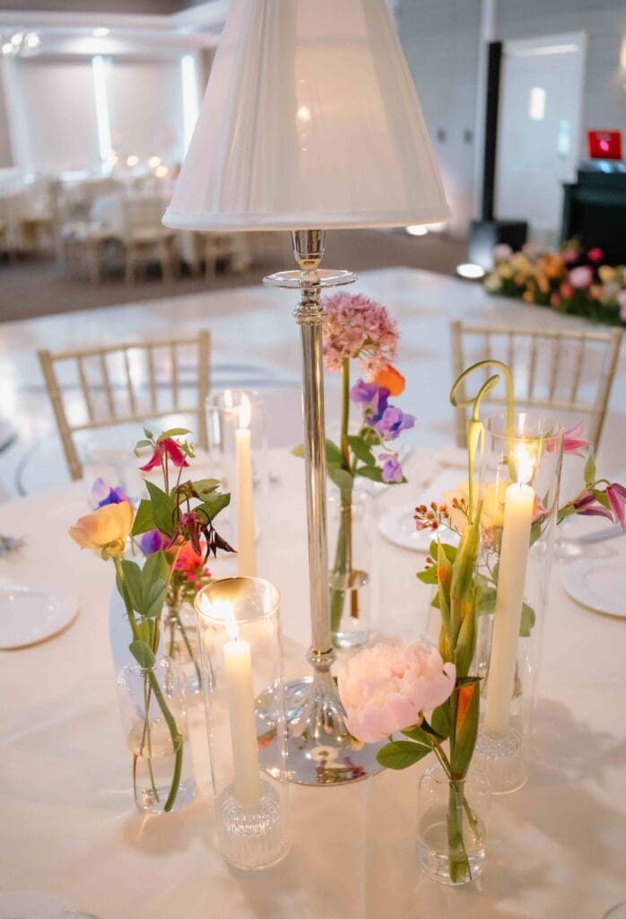 Modern Cape Cod wedding reception setup featuring glass candlesticks, delicate bud vases, and a polished lamp centerpiece surrounded by seasonal flowers.