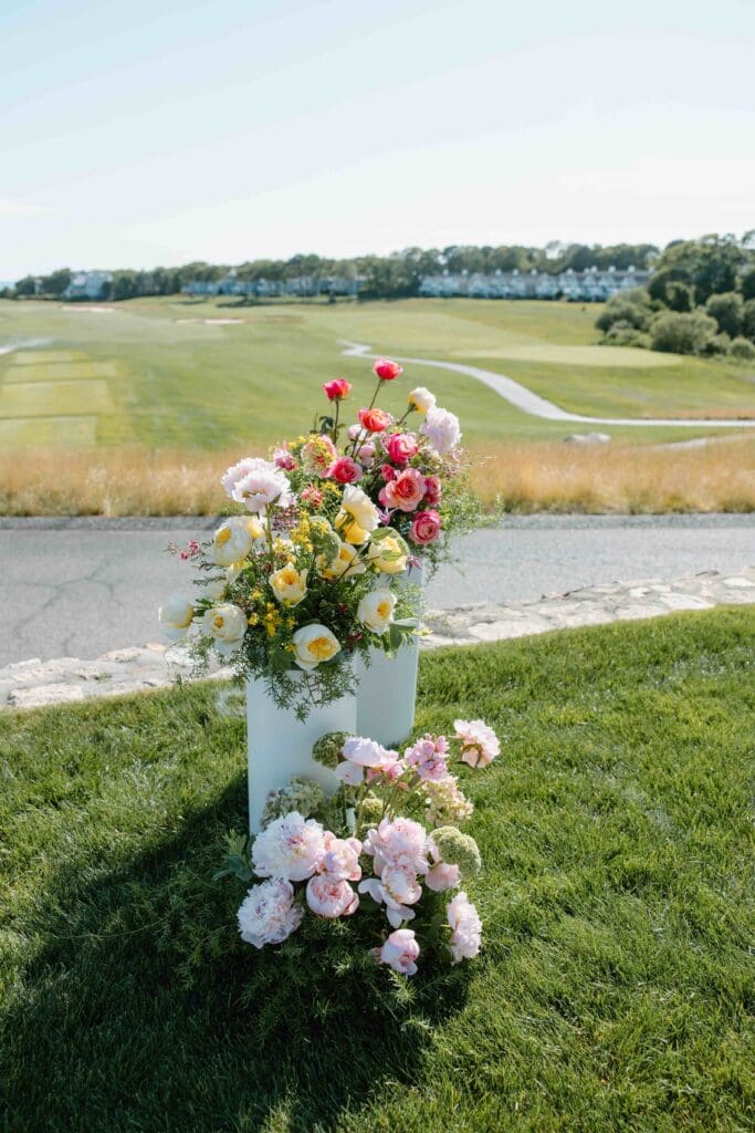 Close-up of vibrant pink, yellow, and coral spring flowers arranged on sleek white columns with the coastal horizon behind them at New Seabury Country Club.