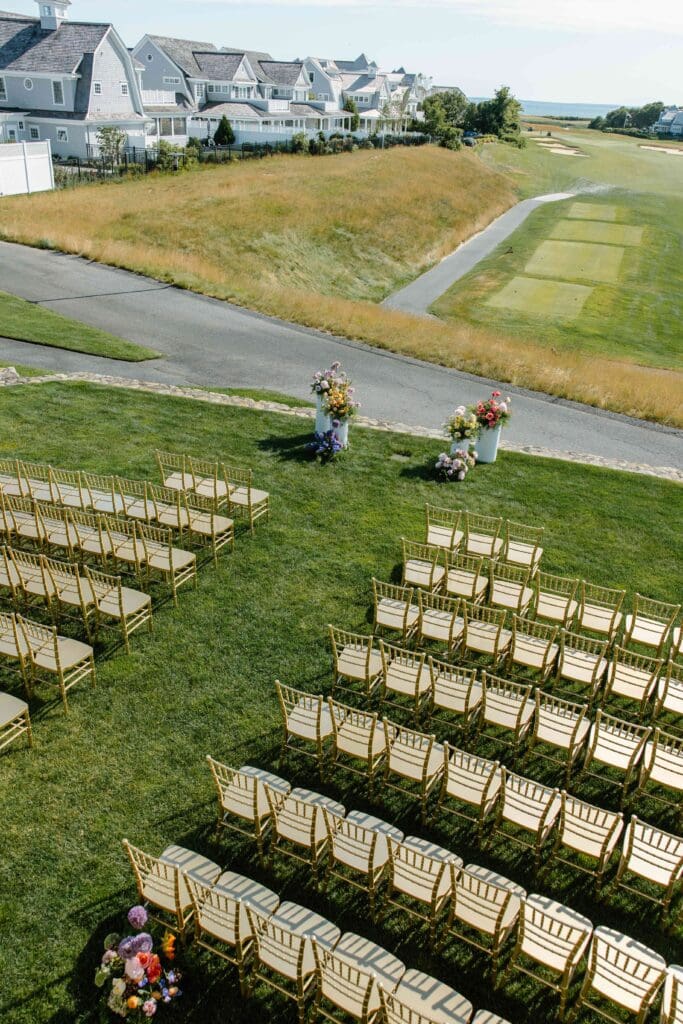 Aerial view of the outdoor ceremony setup at New Seabury Country Club in Cape Cod, featuring rows of gold chairs and modern white pedestals filled with colorful seasonal flowers overlooking the golf course and ocean horizon.