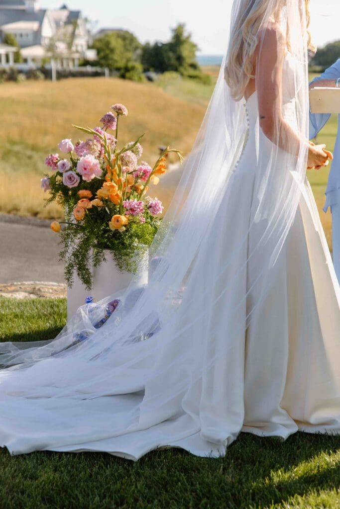 Bride’s veil and gown flowing beside bright coral and yellow floral arrangements during a sunny Cape Cod ceremony at New Seabury Country Club.