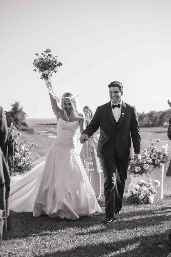 Black-and-white photo of newlyweds celebrating down the aisle, bride lifting her modern bouquet amid guests at New Seabury Country Club.