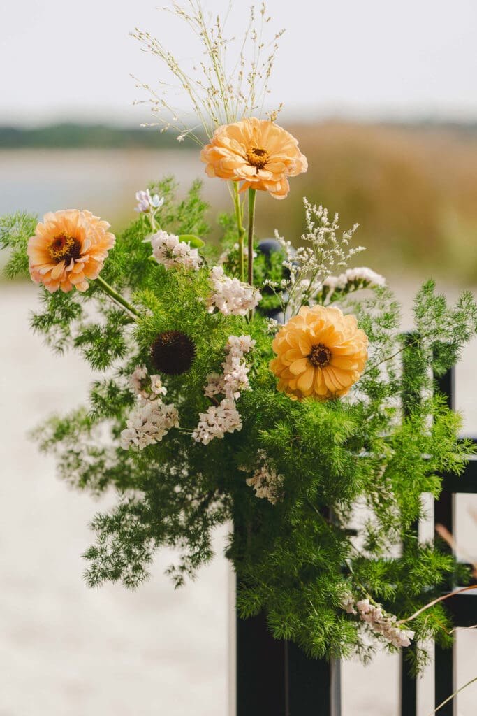 Close-up of a golden-peach zinnia and delicate coastal greenery arranged against the backdrop of Cohasset’s shoreline.