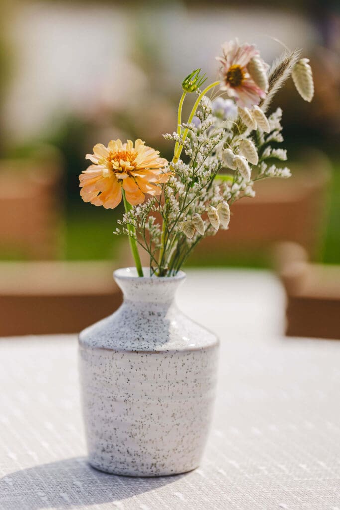 Minimal bud vase with a single peach zinnia and wispy greenery styled on a soft green runner at a coastal backyard wedding.