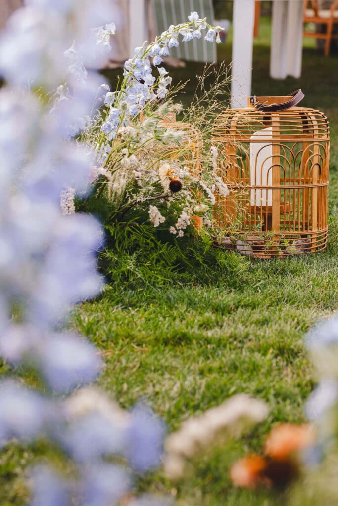 Meadow-style floral installation with blue delphinium, grasses, and woven lanterns placed along the lawn for a coastal backyard ceremony.