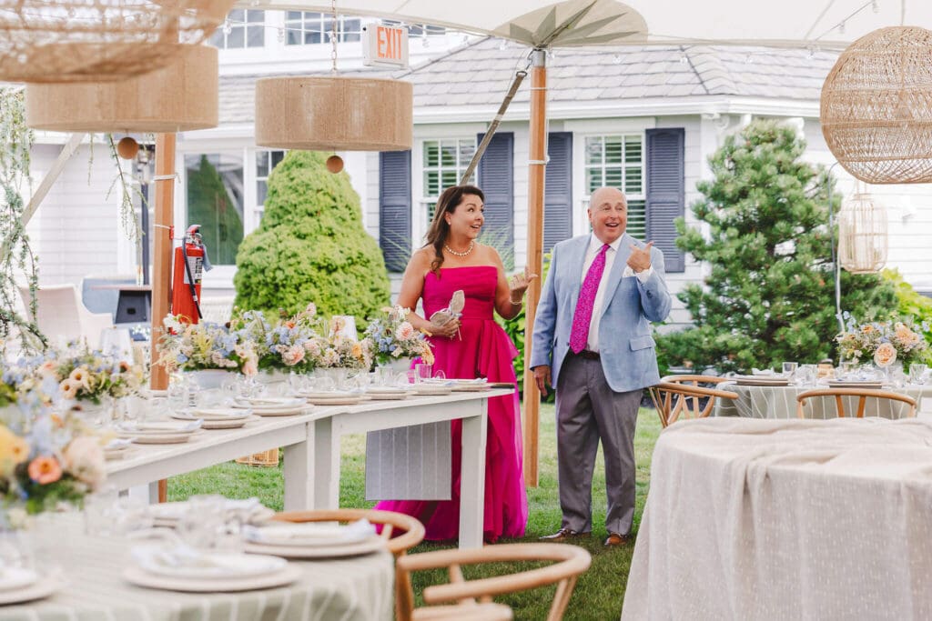 Guests walking through the tented reception setup featuring rattan pendant lights, long tables, and coastal-inspired floral centerpieces at an at-home Cohasset wedding.