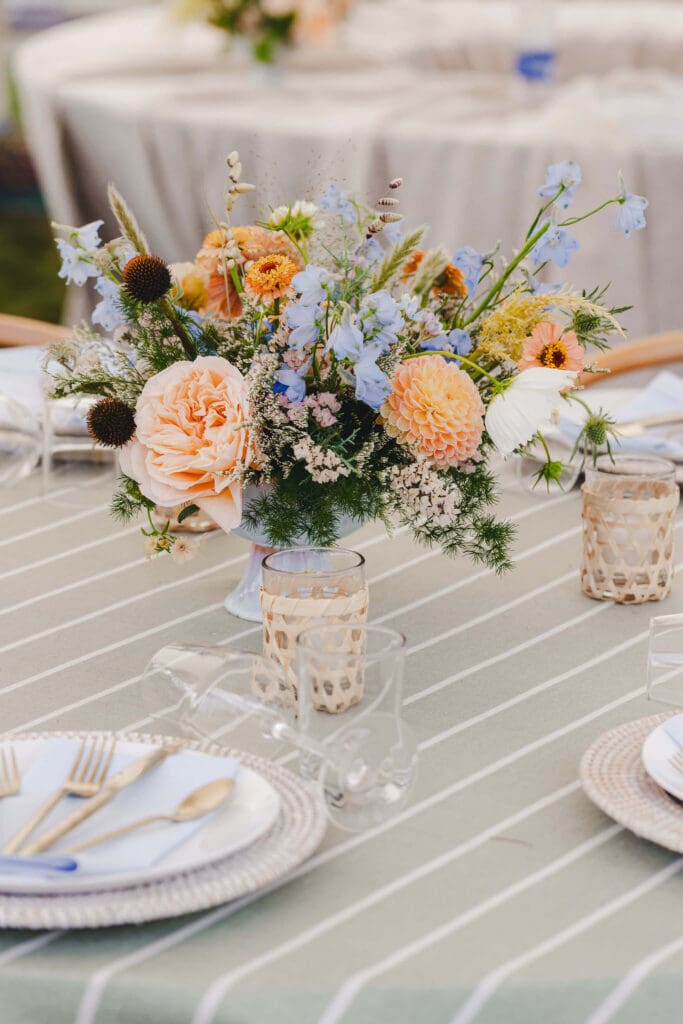 Garden-style compote centerpiece of peach zinnias, dahlias, blue blooms, and coastal greenery set on a striped linen table with woven accents.