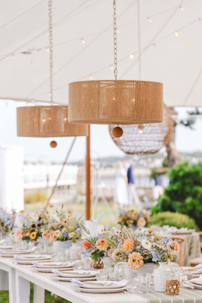 Rattan pendant lights hanging under a sailcloth tent above long white reception tables lined with airy coastal floral arrangements in peach, blue, and soft yellow.