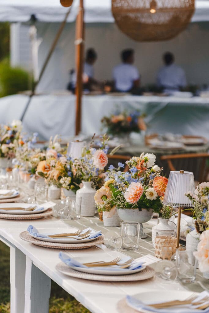 Close-up of long reception tables with compote arrangements of golden-peach zinnias, blue delphinium, and soft grasses, styled beneath rattan pendants at a casual coastal backyard wedding.