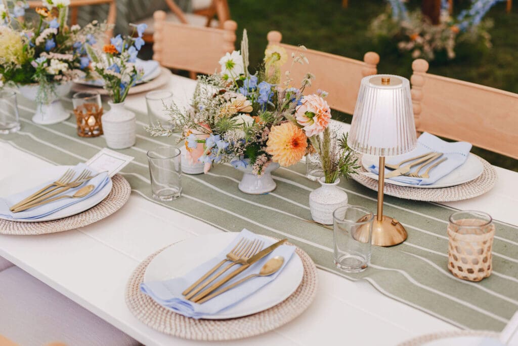 A long white reception table set with green striped table runners, bud vases, coastal garden-style centerpieces, woven chargers, and soft blue napkins, surrounded by wooden spindle-back chairs.