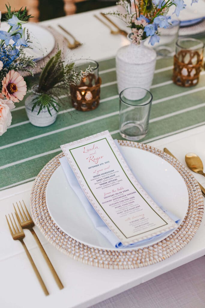 Close-up of a place setting featuring a textured charger, pale blue napkin, printed menu, gold flatware, and surrounding bud vases with single stems and grasses.