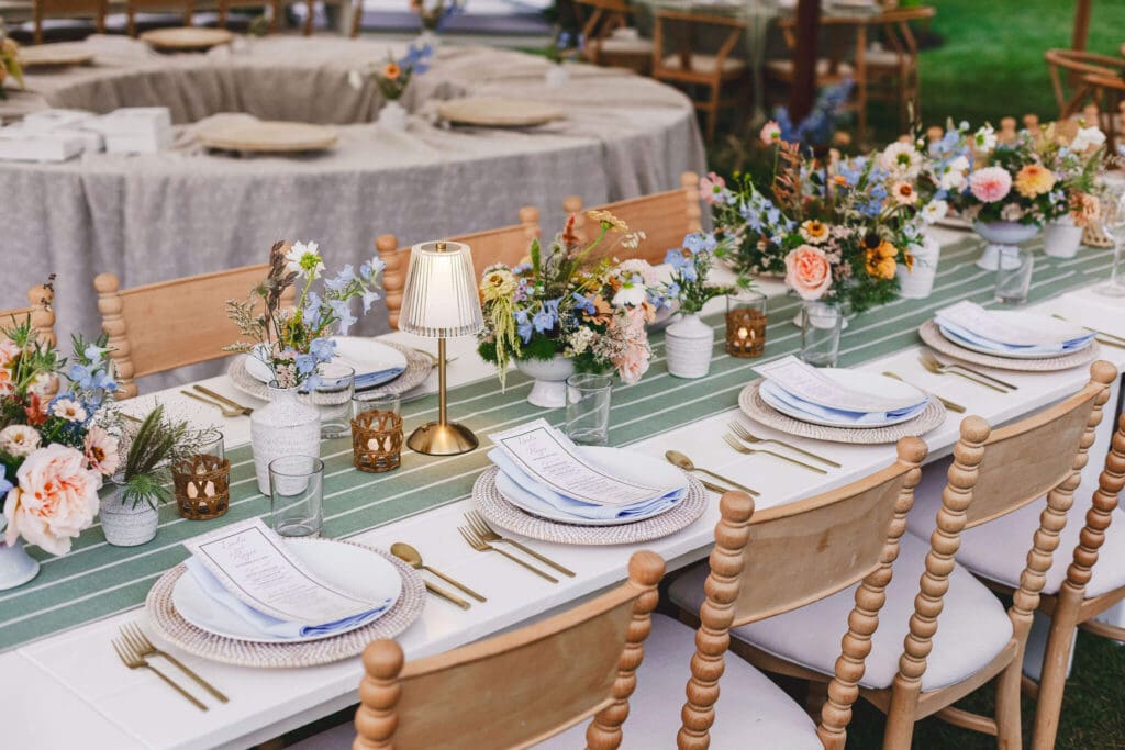 A long dinner table under a tent, styled with layered coastal florals in muted peaches and blues, glassware, white plates, and woven chargers, with the couple’s backyard home blurred softly in the background.