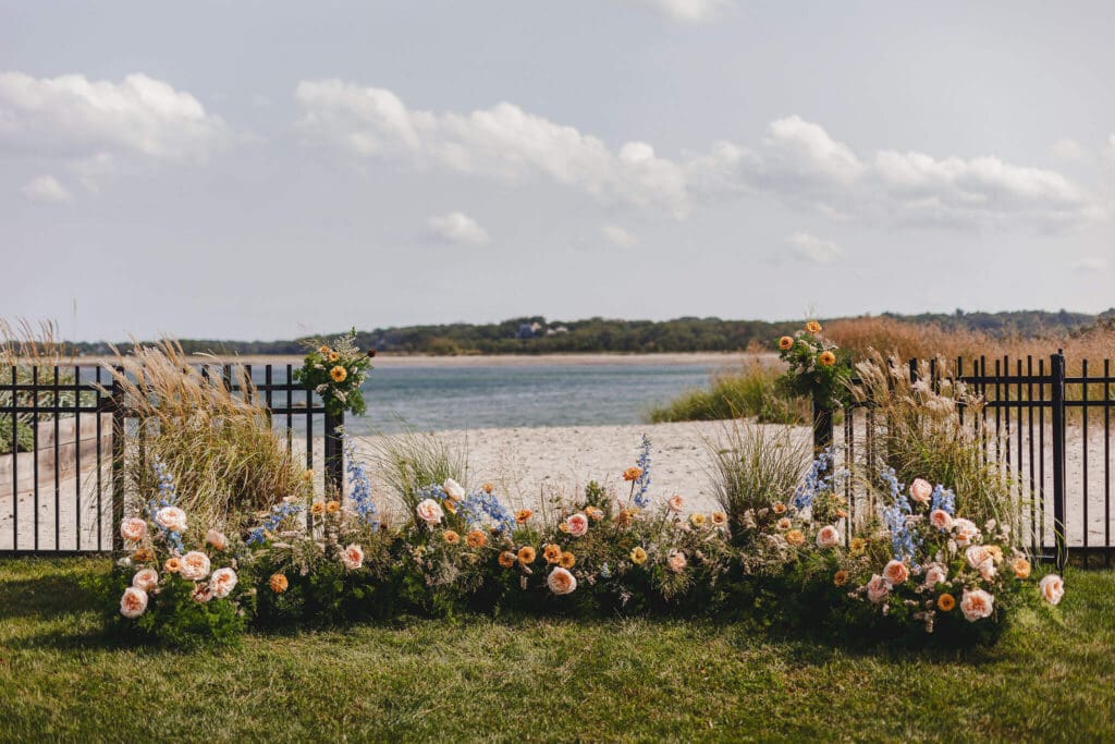 Ground-level coastal ceremony installation featuring peach zinnias, blue delphinium, grasses, and meadow-style florals framing the ocean view in Cohasset.