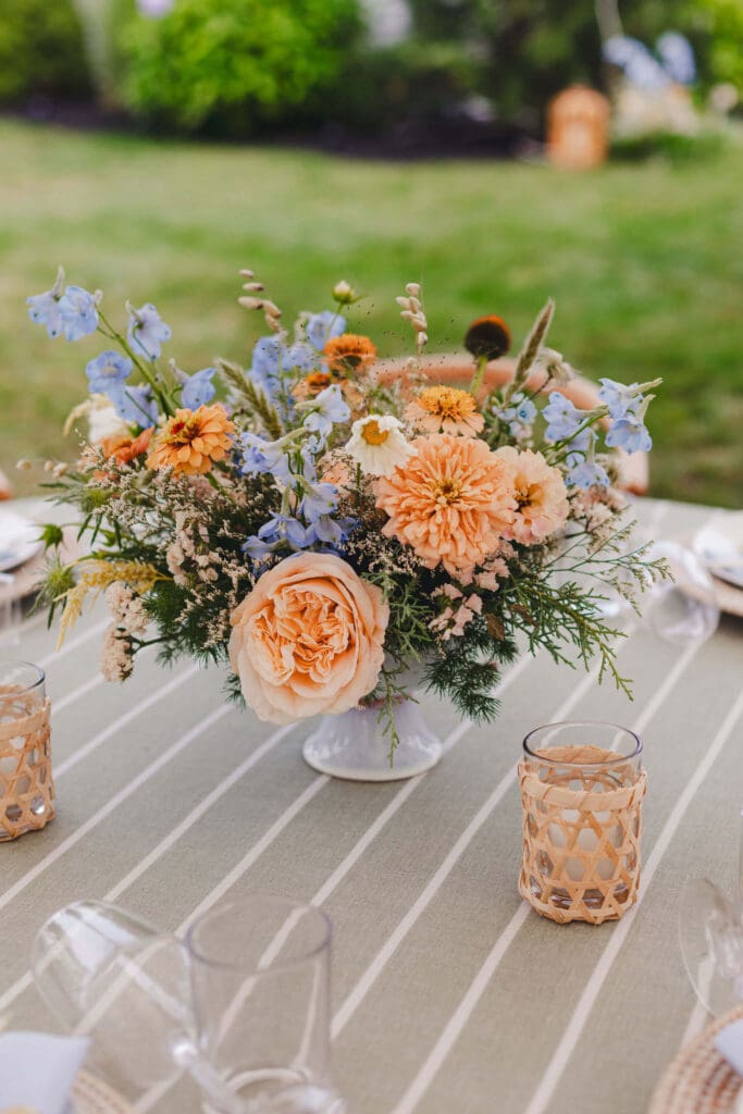 Soft, garden-style centerpiece of golden zinnias, dahlias, cosmos, and blue delphinium on a striped linen table at a coastal backyard wedding.
