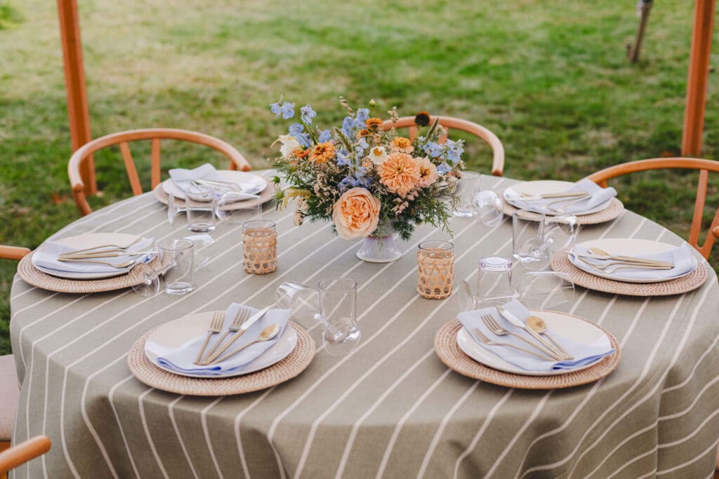 Round reception table with striped linen, woven chargers, gold flatware, and a peach-and-blue coastal centerpiece under a backyard tent.